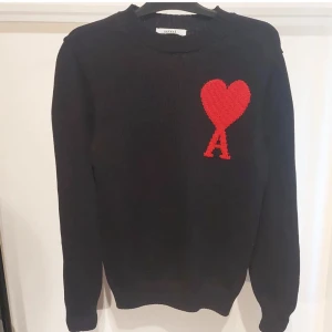 Svart stickad tröja med rött hjärta från Ami Paris - Svart stickad tröja från Ami Paris med ett stort rött hjärta och bokstaven A broderat på bröstet. Klassisk rund halsringning och långa ärmar. Perfekt för dig som gillar stilrena plagg med en twist.