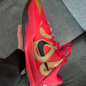 Nike Kobe V Protro University Red/Black - Säljer ett par Nike Kobe V Protro i färgen University Red/Black med guldiga detaljer och svart sula. Skorna har snörning, svart swoosh och en cool guldig design på sidan. Perfekta för dig som gillar basket och vill sticka ut med färgstarka sneakers. Kobe 5, helt oanvända. Allt og ingår