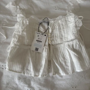 Vit spetsprydd croppad blus - Säljer en söt vit croppad blus i st XS med spetsdetaljer och knytband från BERSHKA. Endast testad, prislapp finns kvar