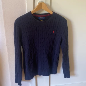 Mörkblå kabelstickad tröja från Polo Ralph Lauren - Mörkblå kabelstickad tröja från Polo Ralph Lauren med rund halsringning och röd broderad logga på bröstet. Klassisk modell med långa ärmar och ribbade muddar. Perfekt för en stilren och tidlös look.