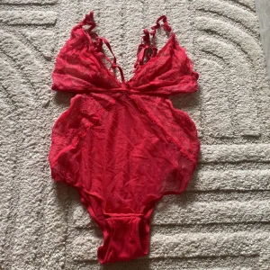 Röd spetsig bodysuit med justerbara axelband - Säljer en röd bodysuit med spetsdetaljer och tunna, justerbara axelband. Bodyn har v-ringning och transparent spets framtill samt släta partier baktill. Perfekt för att lyfta din outfit med en touch av färg och elegans.