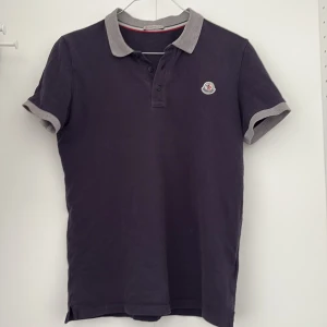 Mörkblå Moncler pikétröja med grå krage - Säljer en mörkblå pikétröja från Moncler med grå krage och ärmslut. Tröjan har knappslå framtill och Moncler-logga på bröstet. Perfekt för en stilren och sportig look.