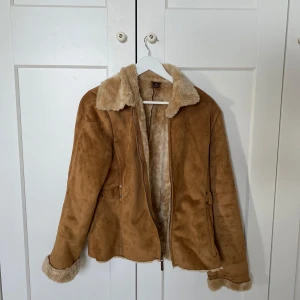 Brun teddyjacka med fuskpälsfoder - Säljer en brun teddyjacka med mjukt fuskpälsfoder och krage. Jackan har dragkedja framtill och pälsdetaljer vid ärmslut och krage. Perfekt för kyligare dagar och ger en snygg retrovibe. Sitter som en S/M