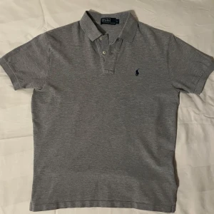 Grå pikétröja från Ralph Lauren - Säljer en klassisk grå pikétröja från Ralph Lauren med korta ärmar och den ikoniska loggan broderad på bröstet. Perfekt för en stilren och avslappnad look. Väldigt snygg Piké som passar formella evenemang men även vardagligt bruk! 