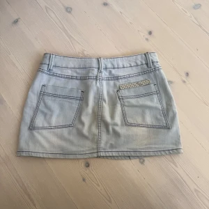 lika jeanskjol med strasstenar - Säljer en så unik jeanskjol med lila kontrastsömmar och fickor både fram och bak. På ena bakfickan finns en detalj med glittriga strasstenar. Köpt second hand. Står tyvärr ingen storlek men skulle uppskatta storlek M