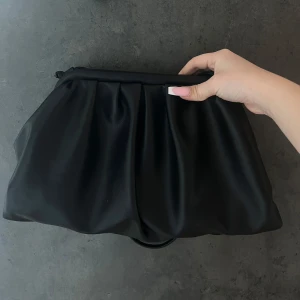 Svart clutch - Trendig svart axelväska med mjuk, puffig form och veckad design. Väskan har en minimalistisk look och stängs med dragkedja upptill. Perfekt accessoar för att lyfta din outfit.