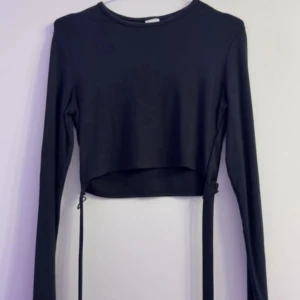 Svart croppad långärmad topp från H&M - Säljer en svart croppad långärmad topp från H&M med knytband i midjan. Enkel och stilren design som passar perfekt till jeans eller kjol. Mjuk och stretchig bomullskvalitet för en bekväm känsla.