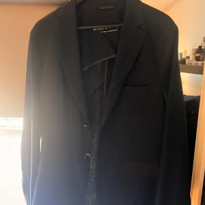 Mörkblå kavaj från Bruun & Stengade - Säljer en stilren mörkblå kavaj gjord på linne från Bruun & Stengade i klassisk passform. Kavajen har två knappar framtill, klassiska slag och fickor med lock. Perfekt för dig som vill ha en tidlös och elegant look. Använd en gång och har inga tecken på användning. Hör av er vid minsta fundering!