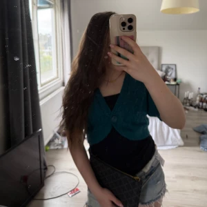 Turkos glittrig croppad väst - Säljer en turkos glittrig väst med kort ärm och croppad passform. Västen har v-ringning och knäpps framtill med en knapp. Perfekt för att addera lite färg och glitter till din outfit.