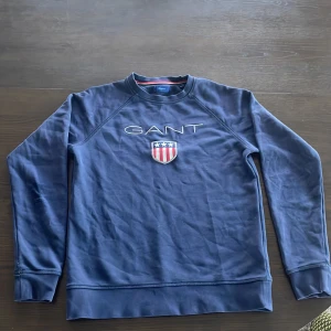 Blå sweatshirt från GANT - Mörkblå sweatshirt från GANT med broderad logga och sköld på bröstet. Klassisk rund halsringning och långa ärmar. Perfekt för en avslappnad och stilren look. SLITNA ARMBÅGAR 