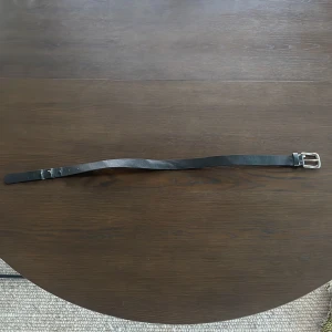 Svart skärp med silverfärgat spänne - Säljer ett klassiskt svart bälte med silverfärgat spänne. Bältet är tillverkat i skinn och har en enkel, stilren design som passar till många olika outfits. Perfekt för dig som gillar minimalistiska accessoarer.