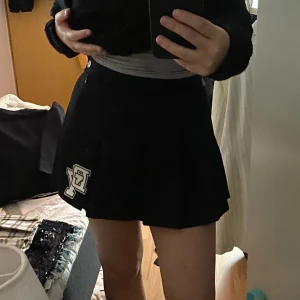 Svart tennis kjol - Svart tennis kjol med bokstäverna ”DY” på framsidan. Plisserad. Blixtlås på sidan med knapp också. Sitter fint! Är 163cm.