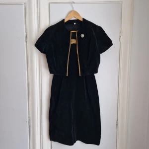 VINTAGE klänning i sammet (med jacka) - Helt fantastiskt tjusig vintageklänning i tät och tung svart sammet. Hög kvalitet och mycket gott skick!  Dragkedja i den v-ringade ryggen. Tillhörande jacka. Silkes-aktiga detaljer i grön-guldig kulör. Brosch med strass. Passar gissningsvis stl S/36. 