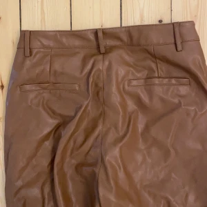 Bruna utsvängda skinnbyxor Ginatricot - Utsvängda bruna skinnbyxor från ginatricot. Är något croppade vid anklarna. Storlek 38. Normal användning, inget synligt slitage