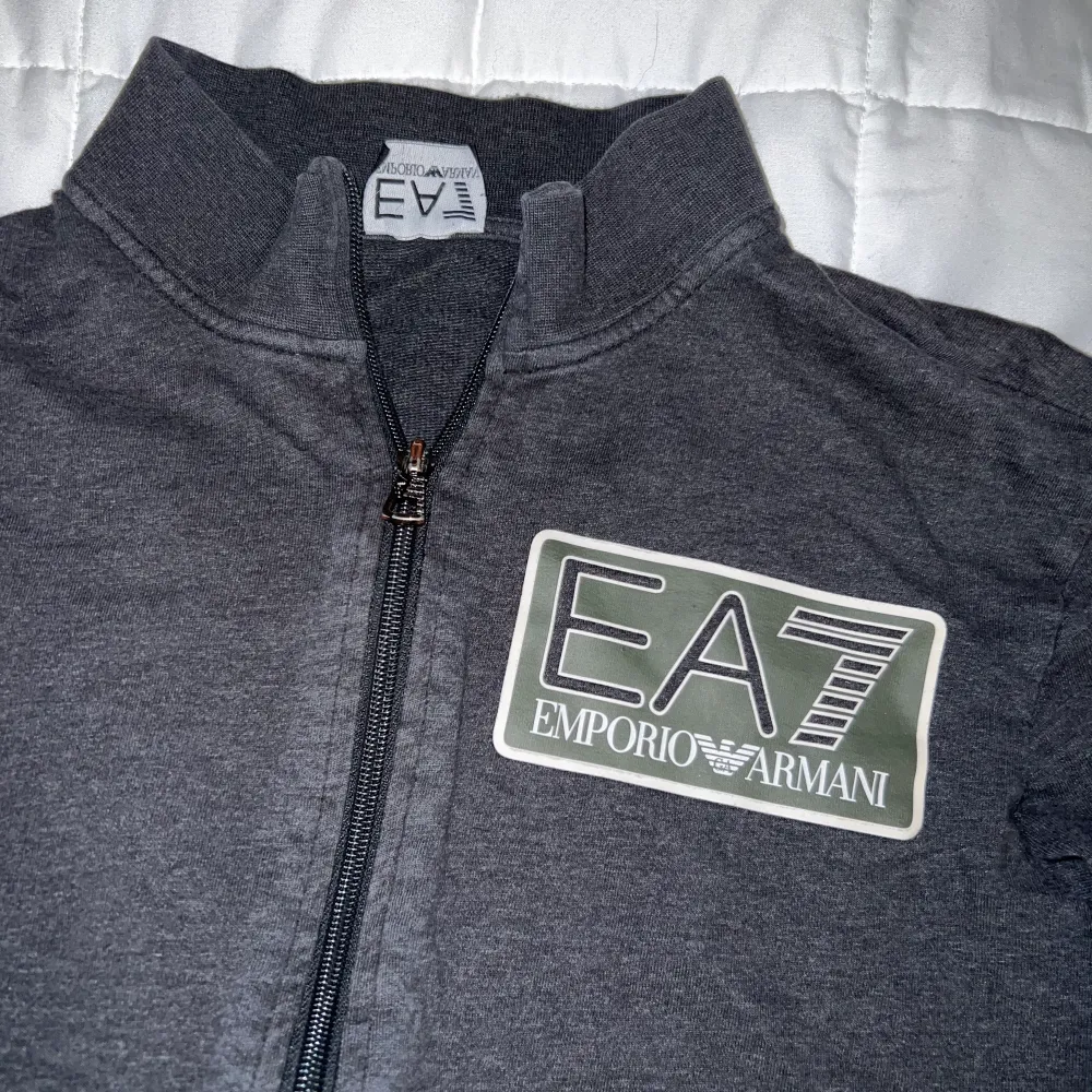 En fin mörkgrå Armani (ea7) tröja med zipper. Märket är i reflex och är delförklaring svår att få en bra bild på tröjan i helhet. Men de är mörkgrön färg i märket också. Storlek M. Hoodies.