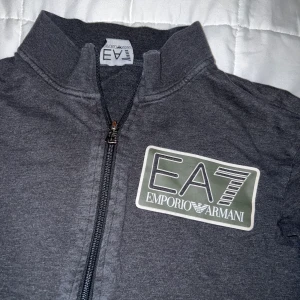 EA7  - En fin mörkgrå Armani (ea7) tröja med zipper. Märket är i reflex och är delförklaring svår att få en bra bild på tröjan i helhet. Men de är mörkgrön färg i märket också. Storlek M