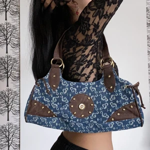 Jeans väska med metall- och läderdetaljer - Rymlig jeans väska i 2000-tals stil. Väskan är köpt på SH och är omärkt. Har använts få gånger efter köpet men har tecken på användning så som slitningar i handtaget finns (se bild nr.3).