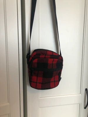 Midjeväska rödrutig  - Skitcool och tuff midjeväska från märket STUHF. Använt några få gånger, säljer för att det ej passade min stil helt enkelt.   Kommer från rökfritt hem 