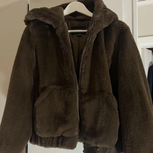 Brun fluffig jacka - Jättemjuk ”fuskpäls", har ett fluffigt intryck med stor luva o ärmar o bred resår i midjan