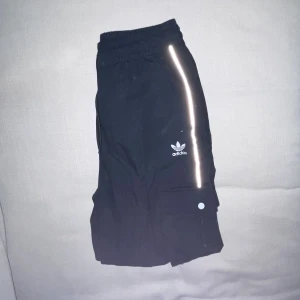 Adidas byxor - Knappt använda Adidasbyxor med reflex längs med benen och två fickor som stängs med tryckknapp. Vanliga fickor vid höften. Resår i midjan och benen.  Nytvättade från ett rökfritt hem, pälsdjur finns men inte på kläderna.  250kr eller bud.