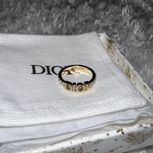 Dior ring - Säljer min äkta Dior ring. En av Diors mest klassiska ringar med guldbelägg, vita kristaller som klär ringens båda sidor samt Dior loggan i mitten. Tyvärr aldrig fått använda den då den är för liten för mig. Presentask, prislapp samt dustbag medföljer
