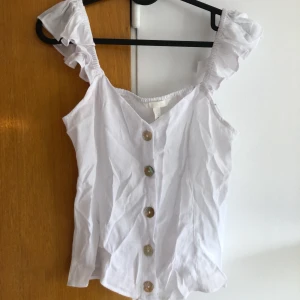 Volangtopp - • Vit volangtopp från H&M • Knappar fram som går att öppna upp och stretchigt material på baksidan • Aldrig använd, prislapp kvar