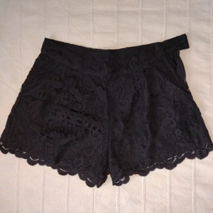 Svarta spetssjhorts - korta shorts med spets  - Svarta korta shorts med spets. Fickor i sidan och shortsen stängs i sidan med dragkedja, hake och knapp. Den vågiga spetskantn går en liten bit nedanför innertyget. Använda en gång och i fint skick. Storlek XS. Yttertyg: 68% bomull och 32% polyamid. Foder