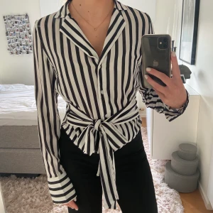 Randig skjorta från NA-KD - Superfin vit- och svartrandig skjorta med knyte i midjan. Storlek: 36. Säljer då den inte kommer till användning. Köparen står för frakten.