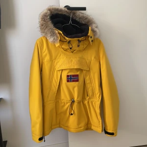 Napapijri vinterjacka - Varm anorak från Napapijri, fin gul färg i Stl XS. Avtagbar pälskant på luvan. Dragkedja på sidan. Dragsko i midjan. Passar jättebra som skidjacka.