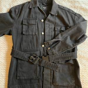 En svart jeansjacka från Bikbok, köptes år 2018, och inte använts sen några år tillbaka. Den är svart/grå i färgen och går ner en bit längs höfterna. Man knäpper den med knappar och ett midjebälte som sitter fast🥳