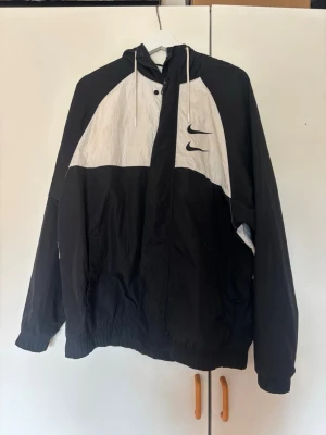 Nike windbreaker/vindjacka - Nike vindjacka med huva, två Swoosh-loggor framtill och stort Nike International/Swoosh-tryck på ryggen.  Sitter även väldigt snyggt oversized på mindre storlekar utan att kännas överdrivet stor.  Bra begagnat skick och tvättas innan den skickas 