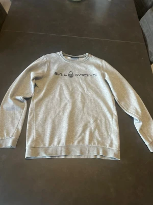 Grå sweatshirt från Sail Racing - Snygg grå sweatshirt från Sail Racing med rund halsringning och svart logotyptryck på bröstet. Tröjan har långa ärmar, ribbade muddar vid ärmslut och nederkant samt en liten logga i nacken. Perfekt till en avslappnad stil. Men på båda armbågarna så är den lite sliten.