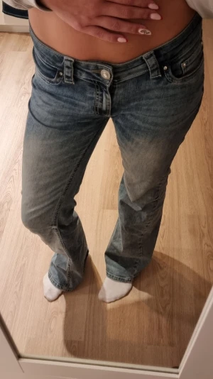 Blå bootcut jeans med fickdetaljer - Säljer ett par blå bootcut jeans med snygg tvätt och slitningar. Jeansen har låg midja, bakfickor med lock och knapp. Perfekta för en avslappnad och trendig llook. Lite slitna längst ner.