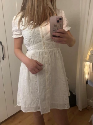 Vit broderad klänning - Söt vit kort klänning med spetsdetaljer och knappar framtill. Klänningen har korta puffärmar och markerad midja som ger en feminin siluett. Perfekt för sommarens alla tillfällen och har en romantisk känsla tack vare spetsmönstret. Den är från LTB, men köpt på sellpy.