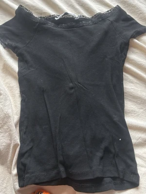 Svart topp med spetskant XS - Säljer en svart topp i storlek XS med snygg spetskant vid halsringningen. Modellen är tight och har korta ärmar, perfekt för att styla med jeans eller kjol. Materialet känns mjukt och stretchigt, vilket gör den bekväm att bära hela dagen.