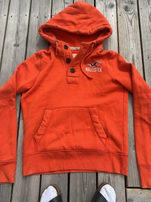 Orange hoodie från Hollister - Snygg orange hoodie från Hollister med broderad logga på bröstet. Hoodien har stor huva, två knappar vid halsen och en klassisk magficka. Perfekt för dig som vill ha en casual och trendig look. Mjuk bomullskänsla och ribbade muddar.