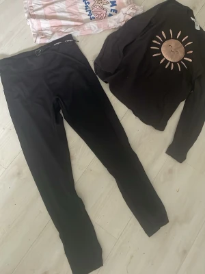 Svart loungewear-set med soltryck - Mysigt loungewear-set med svarta leggings och en svart långärmad tröja med ett soltryck i beige och brunt framtill. Perfekt för chill dagar hemma. Materialet ser mjukt och stretchigt ut för extra komfort.ett litet lagat hål på ett knä