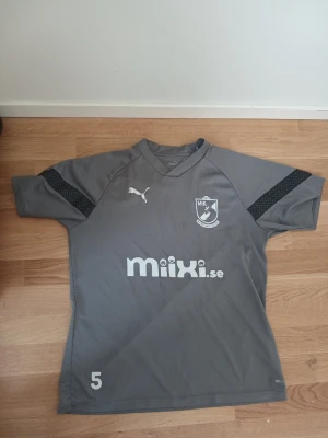 Grå fotbollströja från Puma - Säljer en grå fotbollströja från Puma med svart detalj på ärmarna och klubbmärke på bröstet. Tröjan har tryck med nummer 5 och sponsorlogga på framsidan. Tillverkad i ett lätt och snabbtorkande material, perfekt för träning eller match.