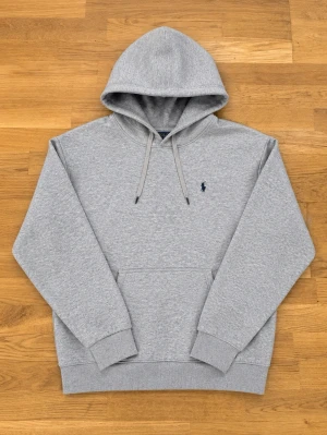 Grå hoodie från Polo Ralph Lauren - Klassisk grå hoodie från Polo Ralph Lauren med huva och dragsnören. Broderad marinblå/svart logga på bröstet och stor känguruficka framtill. Tillverkad i mjuk bomullsblandning och har ribbade muddar vid ärmslut och nederkant. Perfekt för en avslappnad och stilren look.