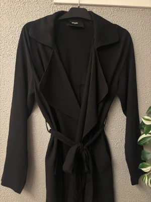 Svart trenchcoat med knytskärp - Stilren svart trenchcoat med långa ärmar och brett slag. Knytskärp i midjan för en snygg siluett och öppen front. Tillverkad i ett mjukt, tunnt tyg som ger ett avslappnat intryck. Perfekt för lager på lager och enkel att matcha.
