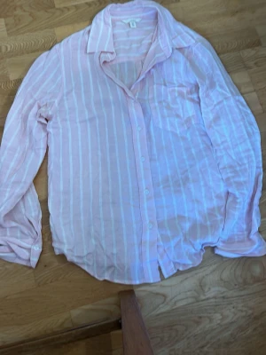 Rosa randig linneskjorta H&M - Ljusrosa skjorta från H&M i linnemix med vita vertikala ränder. Skjortan har klassisk krage, knappstängning framtill och en bröstficka. Perfekt för en avslappnad och stilren look.
