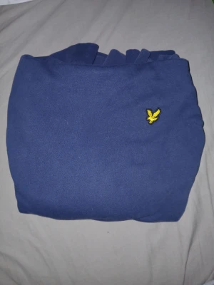 Blå hoodie från Lyle & Scott - Säljer en stilren blå hoodie från Lyle & Scott med klassisk gul logga på bröstet. Hoodien har dragsko i huvan och är gjord i mjukt bomullsmaterial. Perfekt för dig som vill ha en enkel men snygg look.