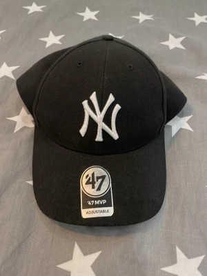Svart New York Yankees keps 47 Brand - Snygg svart keps från 47 Brand med New York Yankees-logga broderad i vitt framtill. Justerbar passform med kardborreband bak där det även står 'Yankees' broderat. Klassisk böjd skärm och ventilerande hål upptill. Perfekt för en sportig och trendig look.
