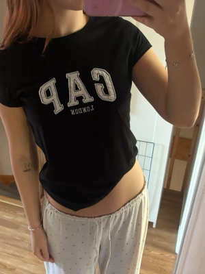 Svart GAP t-shirt med tryck - Snygg svart t-shirt från GAP med vit text 'GAP LONDON' på bröstet. Klassisk passform och rund halsringning. Perfekt till jeans eller shorts för en enkel och stilren look.