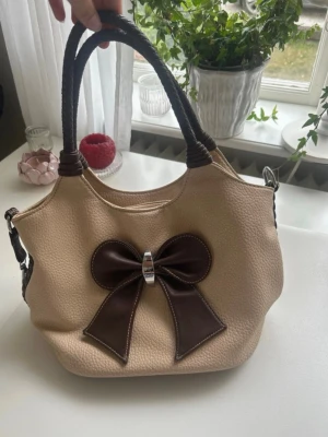 Beige handväska med brun rosett - Snygg beige handväska med en stor mörkbrun rosett framtill och matchande bruna handtag. Väskan har en rymlig insida med dragkedjefack och dekorativt metallspänne på rosetten. Perfekt accessoar för dig som gillar söta detaljer. Tyvvär saknar väskans innerficka piggen till blixtlåset men funkar bra att använda ändå