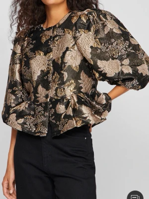Svart & guldig blus från  Vila - Trendig svart blus från Vila med lyxigt jacquardmönster i guld och svart. Blusen har puff & trekvartsärmar, och markerad midja . Unik topp som passar till festliga tillfällen. Den kostade 749kr ny och är endast använd 1 gång, alltså är den som ny i skicket.