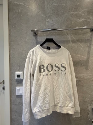 Vit sweatshirt från Hugo Boss - Vit sweatshirt med stor BOSS-logga och texten HUGO BOSS framtill. Tröjan har rund halsringning, långa ärmar och ribbade muddar vid ärmslut och nederkant. Perfekt för en avslappnad och stilren look.