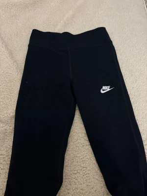 Svarta leggings från Nike - Snygga svarta leggings från Nike med klassisk vit logga på benet. De har hög midja och är tillverkade i ett stretchigt material som sitter skönt på kroppen. Perfekta för träning eller chill dagar. Enkla att matcha med olika outfits.