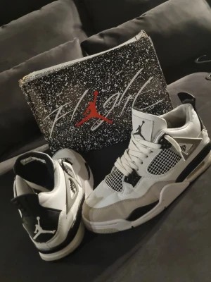 Air Jordan 4 Retro vit/svart/grå - Säljer ett par Air Jordan 4 Retro sneakers i vitt, svart och grått. Skorna har klassisk meshpanel, snörning och ikoniska Jumpman-loggan på plös och häl. Tillverkade i läder och syntetmaterial med detaljer i plast. Box medföljer.