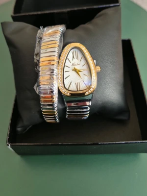 Guld- och silverfärgad ormformad klocka - Unik klocka med ormformad boett och flexibelt armband i guld och silver. Urtavlan är vit med gulddetaljer och romerska siffror, omgiven av gnistrande stenar. Armbandet slingrar sig runt handleden och ger en cool, modern look.